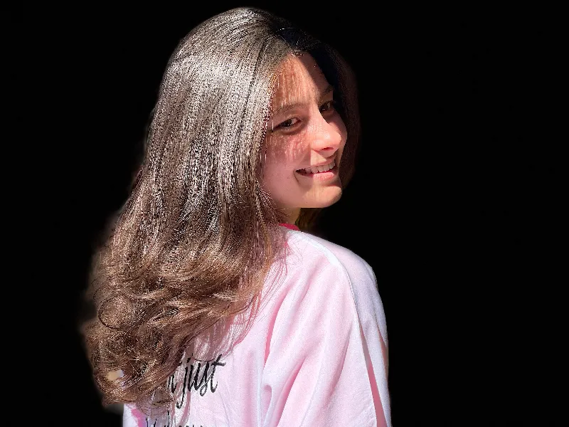 A person with long, shiny brown hair smiles while turning slightly to one side. They are wearing a light pink shirt against a black background, highlighting their hair's texture and volume.