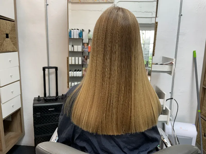 A person with straight, long blonde hair sits in a salon chair, facing a mirror. The salon features shelves with hair products and a hair styling station.