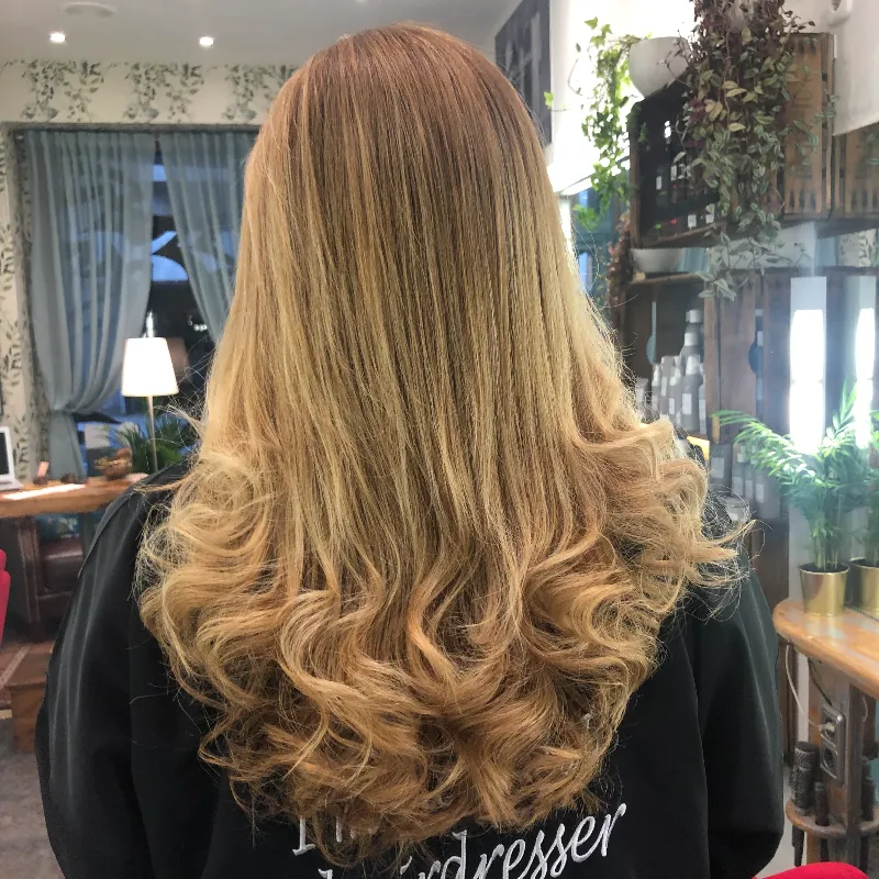 A person with long, wavy blonde hair is shown from the back in a salon setting. The hair is styled with loose curls at the ends. The background features dim lighting, plants, and mirrors.
