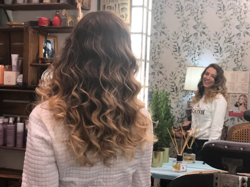 A woman with curly, shoulder-length hair stands with her back to the camera, looking at her reflection in a salon mirror. The room is decorated with floral wallpaper and various hair products. She smiles at herself in the mirror.