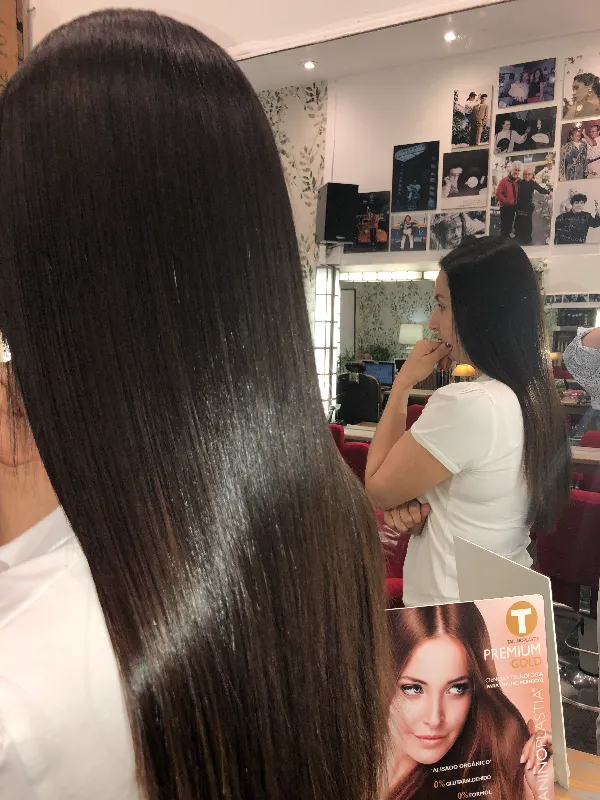 A person with long, straight, shiny dark hair is looking at themselves in a salon mirror. They are holding a magazine or pamphlet. The salon has various hair products and framed pictures displayed on the walls.