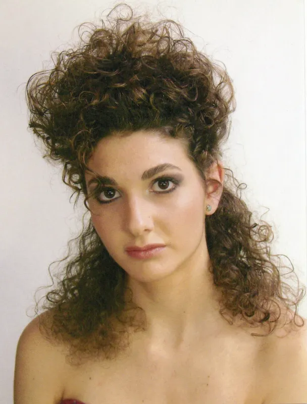 Person with curly, voluminous brown hair styled in a distinctive 1980s fashion, wearing subtle makeup and small earrings, looking at the camera against a plain background.
