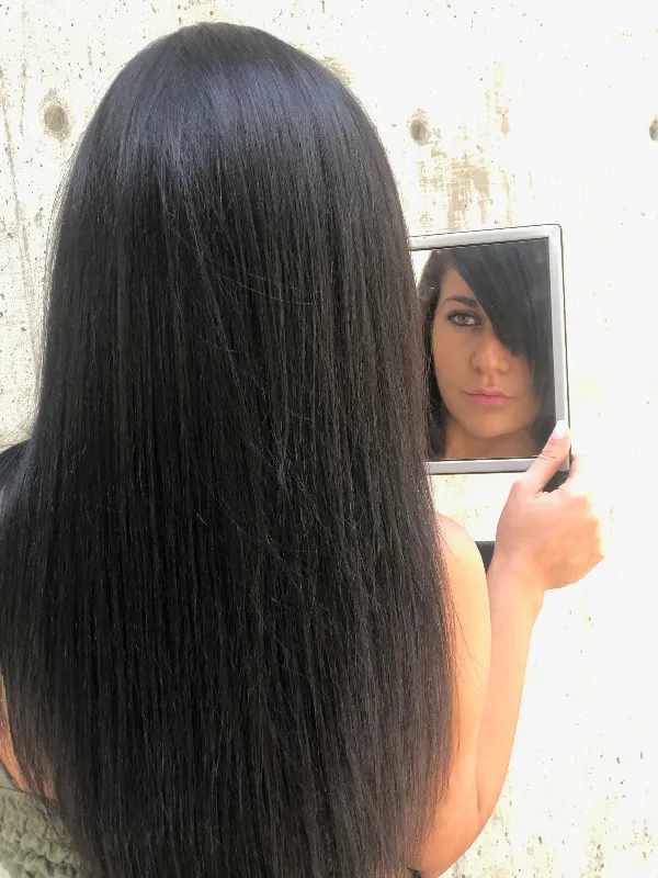 A woman with long, straight dark hair is standing with her back to the camera, holding a rectangular mirror up to her face. Her reflection shows her looking directly into the mirror, set against a plain concrete wall.
