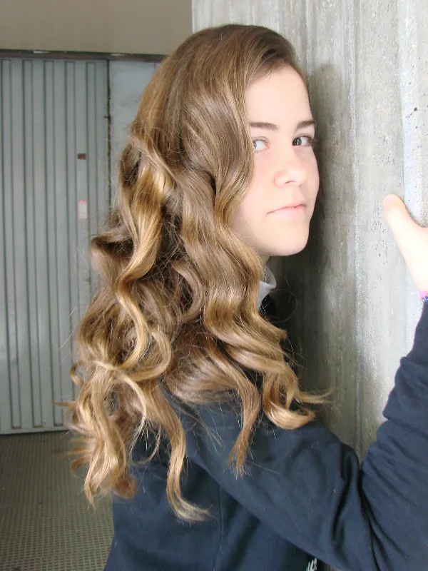 A person with long, wavy brown hair is leaning against a textured wall, gazing at the camera. They're wearing a dark top in a well-lit indoor setting.