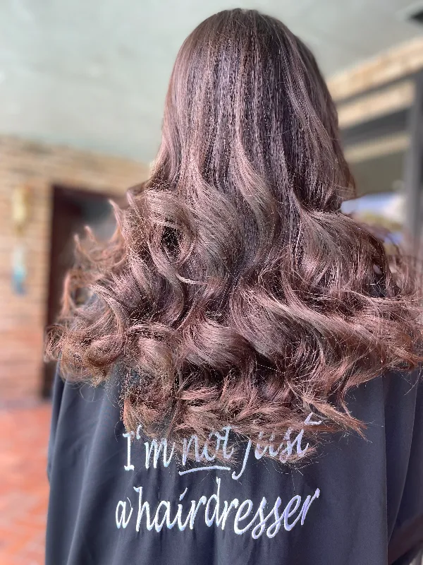 Back view of a person with long, curly brown hair. They are wearing a black shirt with white text that reads, "I'm not just a hairdresser." The setting appears to be indoors with a blurred background.