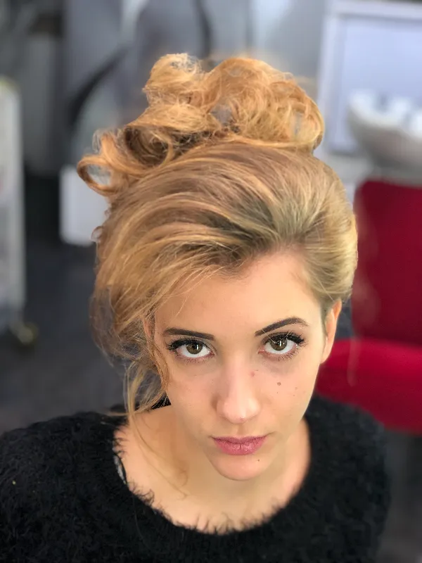A person with light brown, styled hair and a serious expression looks up at the camera. They are wearing a black fuzzy top. The background includes a red chair and blurred salon equipment.