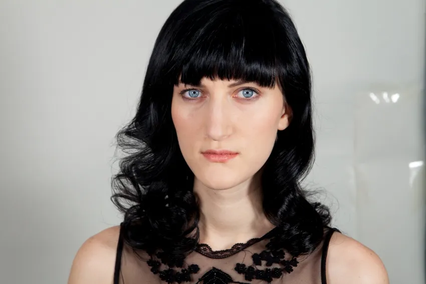 A woman with long, wavy black hair and bangs gazes directly at the camera. She is wearing a black, semi-transparent top with lace details. The background is plain and light-colored.