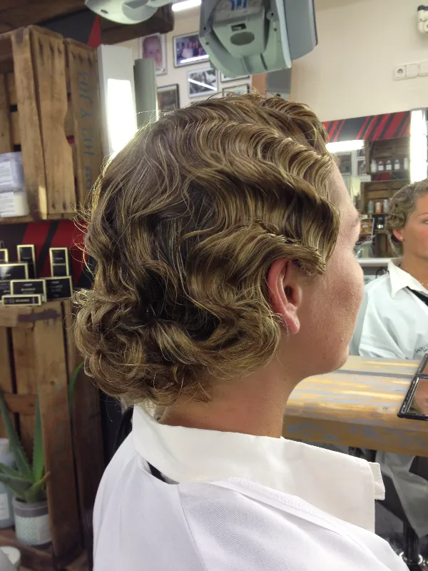 Person with wavy, curled hair sits in a salon chair, reflected in a mirror. They wear a white salon cape, and various hair products are displayed on shelves.