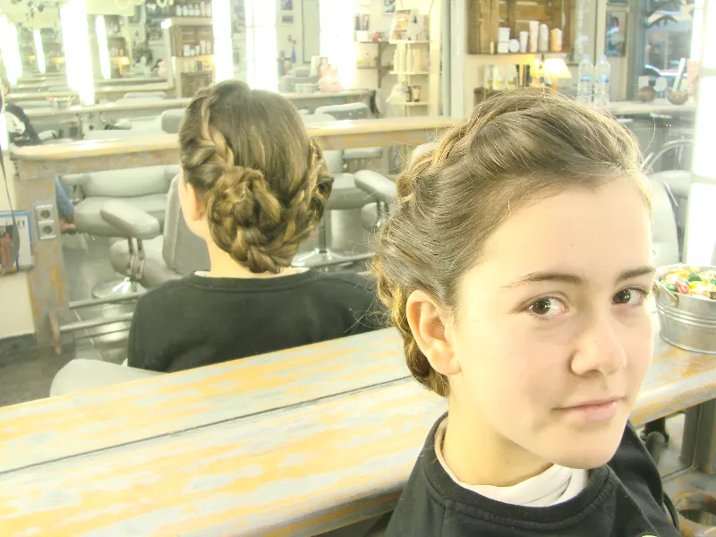 A young girl with braided hair styled in an elegant updo sits in a salon. She looks towards the camera, with mirrors and salon equipment visible in the background.