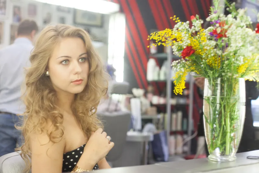 A woman with long, curly hair sits at a salon table with a mirror, wearing a strapless black polka dot top. A tall vase of colorful flowers is placed on the table beside her. The background features salon supplies and a red-striped wall.