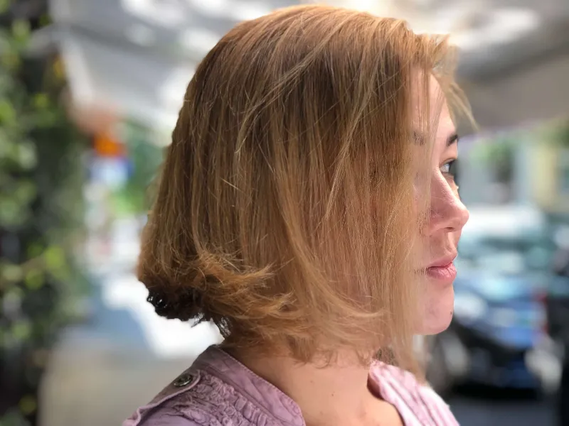 A person with short, light brown hair stands outdoors in profile view, wearing a lavender top. The background is blurred, showing hints of greenery and urban elements.