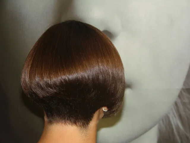 A person with a short, sleek brown bob haircut is facing away from the camera. The haircut is styled with precision, tapering neatly at the nape of the neck. A blurred monochromatic face in the background adds an artistic element.