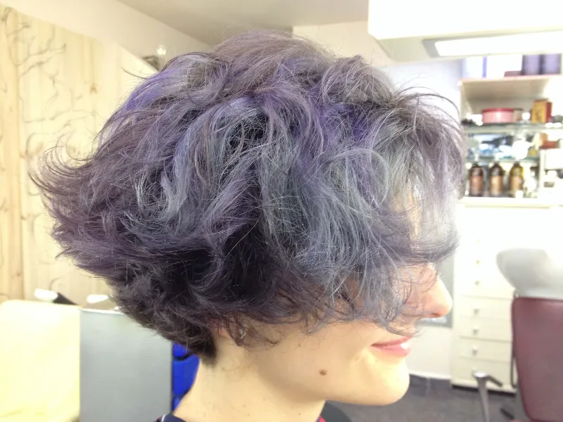 A person with short, wavy hair dyed in shades of purple and blue is sitting in a salon chair. The hair is voluminous and tousled, and the background shows salon equipment and shelves.