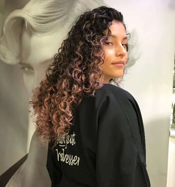 Person with long curly hair and subtle highlights standing in front of a monochrome poster of a vintage hairstyle. They are wearing a dark cape or cover with writing partially visible on the back.