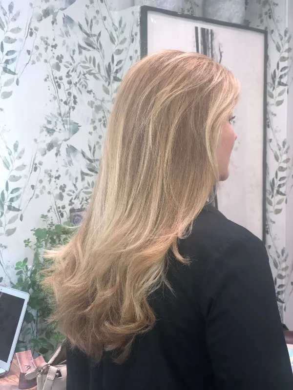 A person with long, layered blonde hair stands in a room with floral-patterned wallpaper. The individual is turned away, showcasing the hairstyle. A table with a laptop and a small plant is visible in the background.