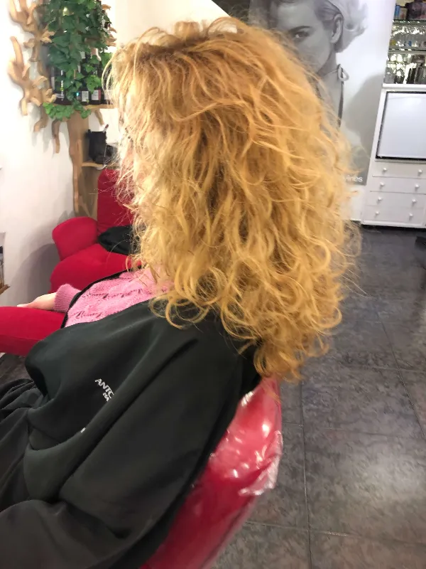 Person with curly, voluminous blonde hair sitting in a red salon chair, wearing a black cape. The background features a poster and salon decor.