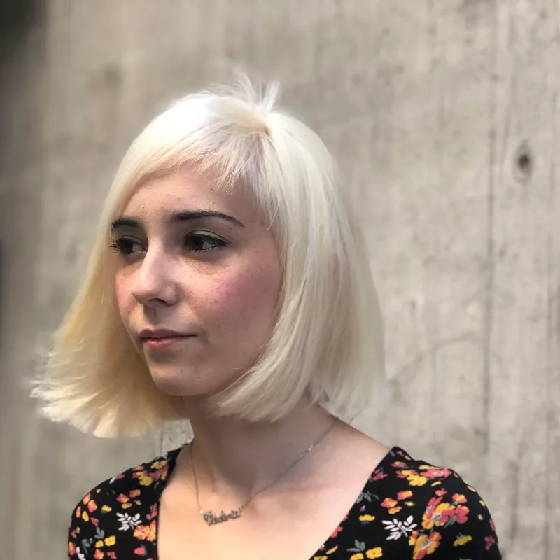 A person with short platinum blonde hair stands against a concrete wall. They are wearing a black shirt with a floral pattern and a necklace. The photo captures a side profile and the expression is neutral.