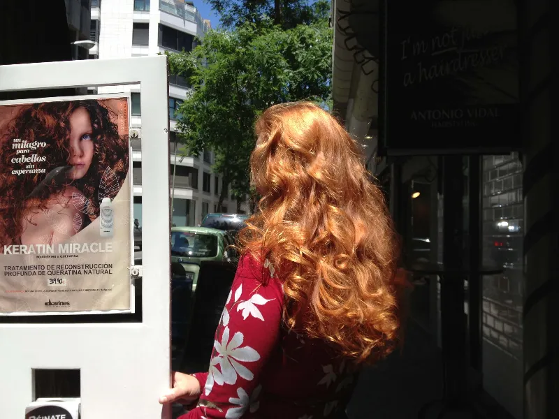 A person with long, curly red hair stands on a city sidewalk, facing away. They are wearing a red floral top. A poster for a hair product is on the left, featuring a model with similar hair. A street and buildings are in the background.