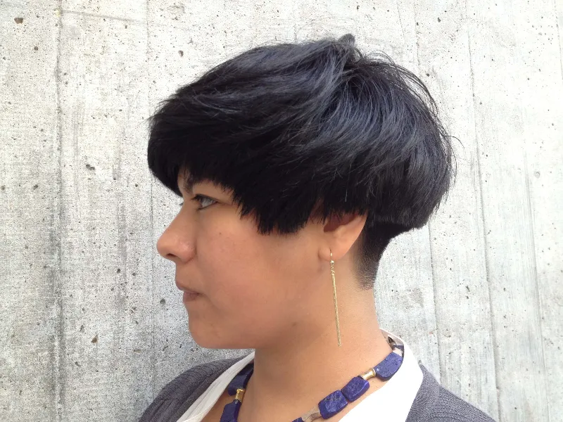 Person with short black hair in a layered style, wearing a white top and a gray cardigan. They have a long, dangling earring and a chunky blue necklace. The background is a textured gray concrete wall, photographed from the side.