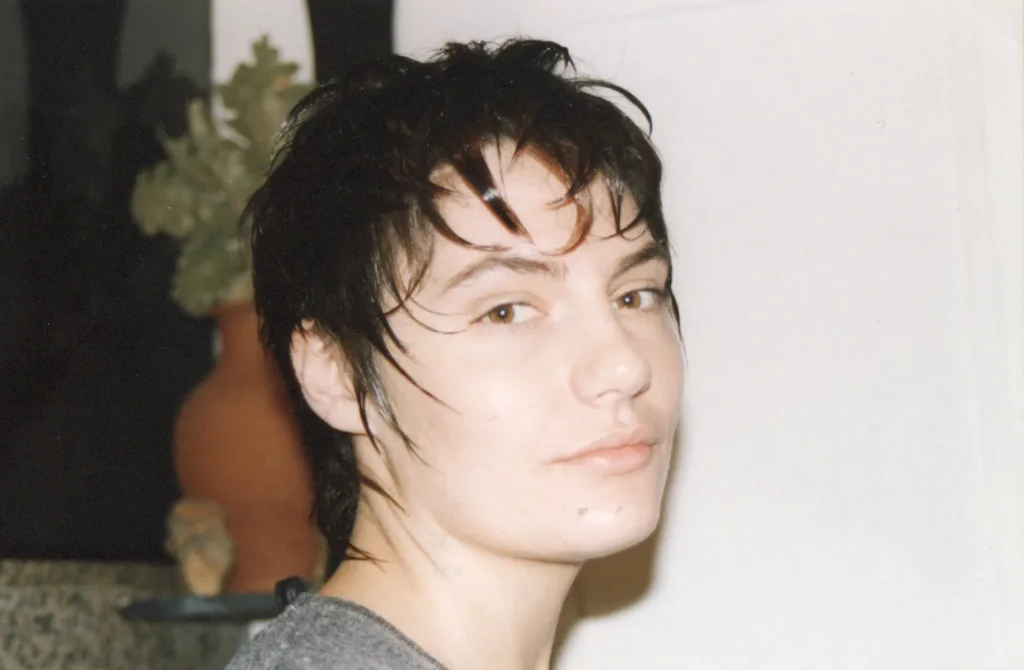 A person with short, wet hair looks towards the camera with a neutral expression. They are indoors, with a terracotta pot and dark foliage in the background. The lighting is soft, highlighting their facial features.
