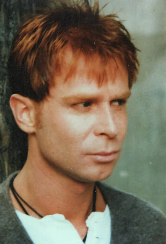 A man with short, reddish-brown hair looks pensively to the side. He is wearing a white shirt with a dark-colored sweater over it and a black necklace. The background is blurred with a hint of a tree trunk.