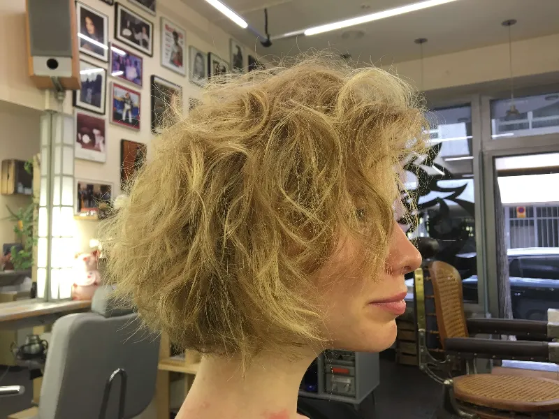 Person with a short, curly blonde hairstyle sitting in a salon. Framed photos are on the wall, and salon chairs are visible in the background.
