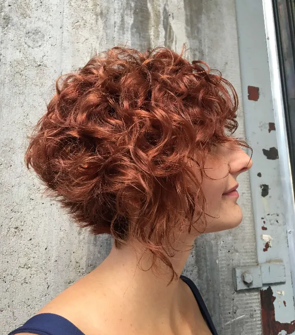 Person with curly, red hair in a short bob hairstyle, facing sideways. The background has a textured, weathered concrete wall and a metal surface. The person is wearing a dark-colored top.