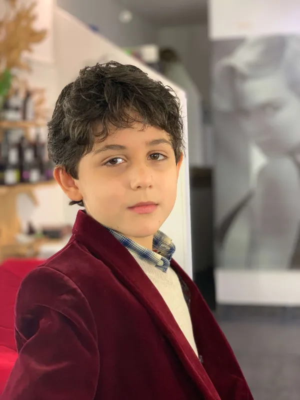 Young child with short dark hair wearing a red velvet jacket and plaid shirt, standing indoors. Blurred background with a large black-and-white photo on the wall. The child looks toward the camera with a neutral expression.