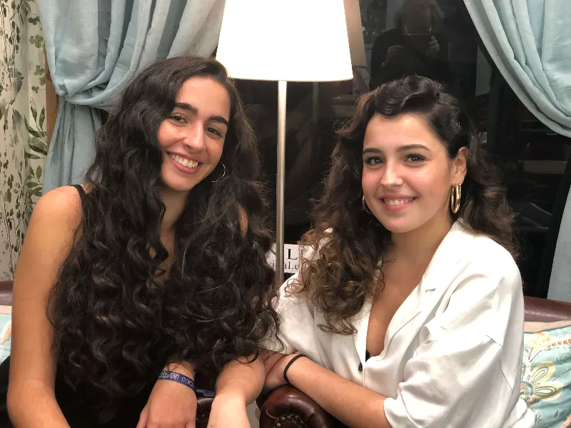 Two women with long, curly hair are sitting indoors in front of a lit lamp and blue curtains. They are smiling at the camera, with one wearing a black dress and the other wearing a white top. They are seated on a leather chair.