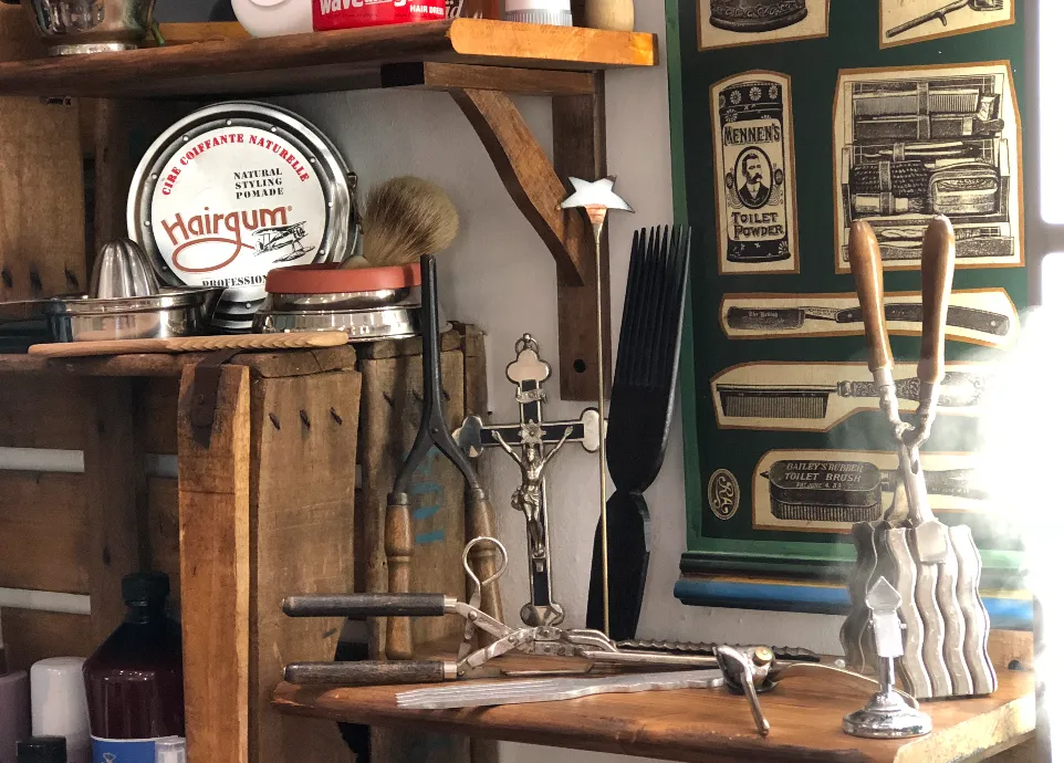 A vintage barber shop interior with antique grooming tools, hair products, and ornate razors displayed on wooden shelves. A decorative wall features nostalgic advertisements and product packaging. Natural light illuminates the cozy space.