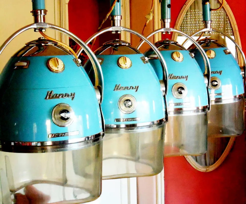 Four vintage blue hair dryers are lined up in a row, attached to a wall with a mirror reflecting them. Each dryer has a chrome finish with the brand name "Henry" displayed on the front. The background is warm-toned.