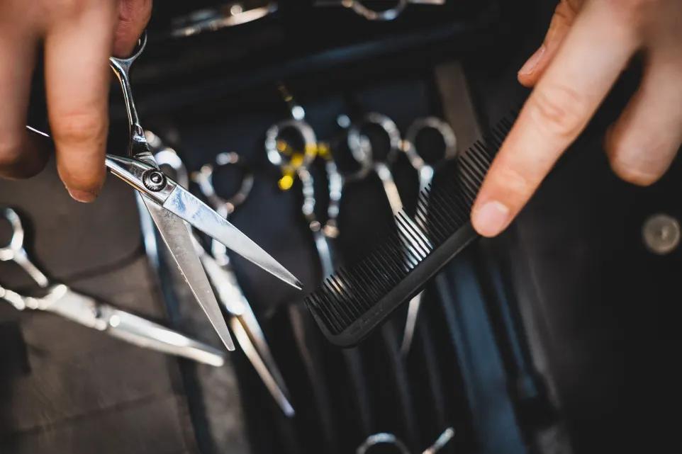 Close-up of hands holding hairdressing scissors and a black comb over a case filled with various hair styling tools. The tools include multiple pairs of scissors and other hair accessories.