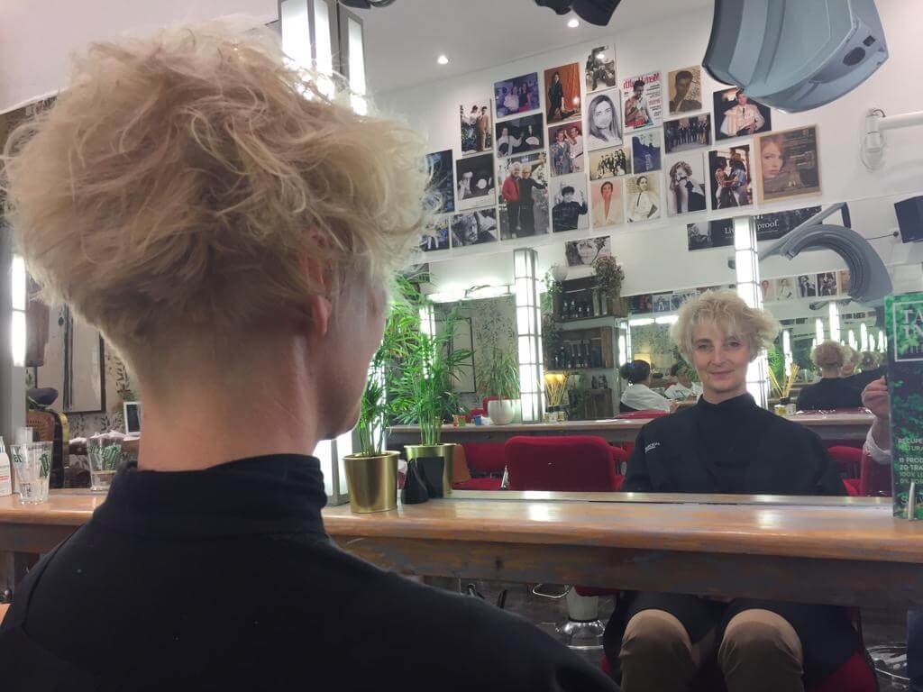 Person with short, curly blonde hair looks at their reflection in a salon mirror. The salon has red chairs, plants, and photos on the wall. The person smiles slightly, wearing a black cape.